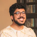 Vitor sorrindo: brasileiro de pele em tons médios, cabelo e barba escuros, óculos de armação grossa, sorrindo em ambiente interno.